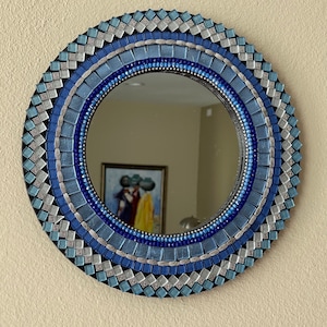 May include: A round mirror with a mosaic frame made of blue and silver tiles. The frame has a geometric pattern and is surrounded by a black border.