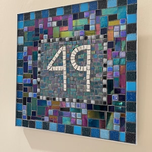May include: A mosaic tile house number sign with the number 49 in white tiles on a black background. The background of the sign is made of small, colorful tiles in shades of blue, green, purple, and black.