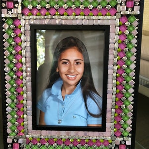 May include: A black picture frame with a mosaic border of pink, green, and silver tiles. The frame is holding a photo of a young woman with long dark hair smiling.