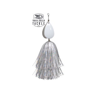 May include: A white and silver fishing lure with a teardrop-shaped blade and a silver, shimmering skirt. The lure has the text "TOOTH SHIELD TACKLE" in a black logo.