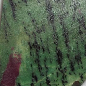 Pode incluir: Uma peça triangular de pedra natural, predominantemente verde com inclusões pretas e vermelho escuro. A pedra tem uma borda áspera e uma superfície polida, com uma veia branca que a atravessa. Dimensões não especificadas.