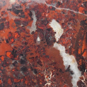 May include: A flat, rectangular slab of polished stone featuring a mottled pattern of deep brick red, dark brown, and black inclusions. A prominent, irregular white vein runs vertically through the center, contrasting with the earthy tones.