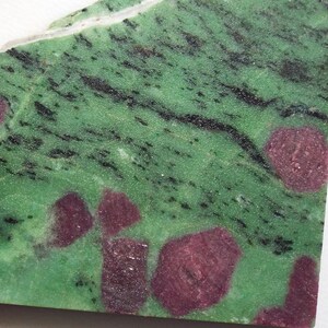 May include: A triangular-shaped piece of zoisite with ruby inclusions. The stone features a vibrant green base with black veining and several deep red ruby spots. A thin white line runs along the top edge.