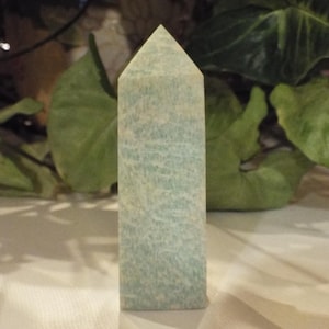 Pode incluir: Uma torre de cristal polido com base hexagonal e topo pontiagudo. A pedra apresenta uma cor azul-esverdeada pálida com padrões brancos, nublados e manchados em toda a sua superfície, exibindo uma textura natural e diversas inclusões minerais.