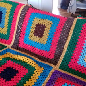 May include: A handmade crochet blanket featuring a repeating granny square pattern in vibrant colors including hot pink, bright yellow, turquoise, dark brown, forest green, navy blue, and purple, joined together with a golden yellow yarn border.