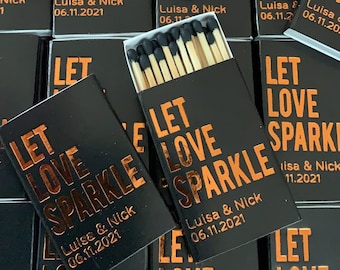 Może przedstawiać: Kolekcja czarnych pudełek z zapałkami z pomarańczowym napisem LET LOVE SPARKLE, Luisa & Nick oraz 06.11.2021. Jedno otwarte pudełko ukazuje drewniane zapałki z czarnymi główkami. Pudełka są ułożone w siatkę, prezentując ten sam nadruk.