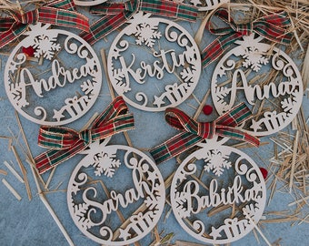 May include: Six wooden Christmas ornaments with red and green plaid ribbon. Each ornament is a circle with a snowflake design and a name written in cursive: Maminka, Laura, Andrea, Kerstin, Anna, and Babicka.