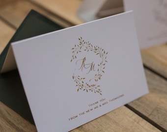 May include: A white thank you card with a gold floral wreath and the initials "R.M." in the center. The text "Thank you from the new Mr. & Mrs. Thompson" is printed below the wreath.