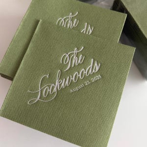 Puede incluir: Servilletas cuadradas verde oliva con texto blanco en cursiva que dice "The Lockwoods" y "August 23, 2025". Las servilletas están apiladas, algunas visibles en embalaje plástico transparente.