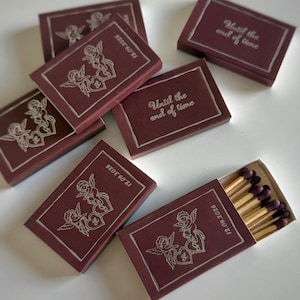 May include: A collection of rectangular matchboxes in deep maroon with silver foil detailing. Some boxes feature a line art illustration of two cherubs with hearts and the date 12.09.2026, while others display the text Until the end of time.
