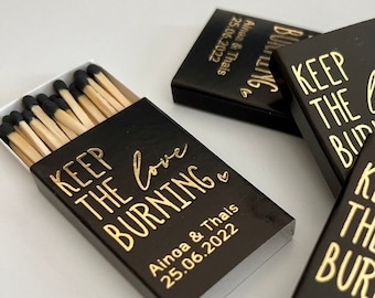 May include: Three black matchboxes with gold foil text that reads KEEP THE LOVE BURNING, Ainoa & Thais, and 25.06.2022. One box is open, revealing wooden matches with black tips. The design includes a small heart icon next to the text.