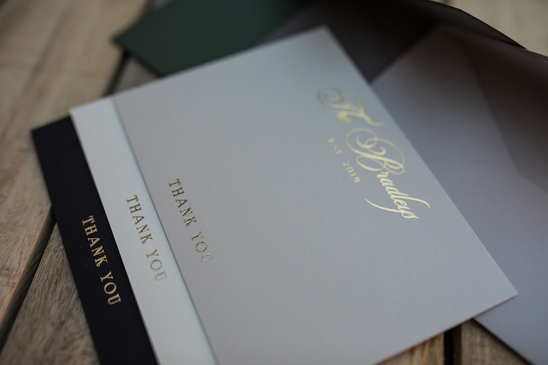 May include: Three thank you cards with gold lettering. The top card is gray with the text "The Bradleys Est 2019" in gold. The middle card is white with the text "Thank You" in gold. The bottom card is black with the text "Thank You" in gold.