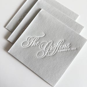 Peut inclure: Une pile de trois serviettes en papier carr&eacute;es gris clair avec une finition textur&eacute;e. La serviette du dessus pr&eacute;sente une calligraphie blanche en relief indiquant The Griffins et la date September 27, 2025, centr&eacute;e sur la surface du papier.