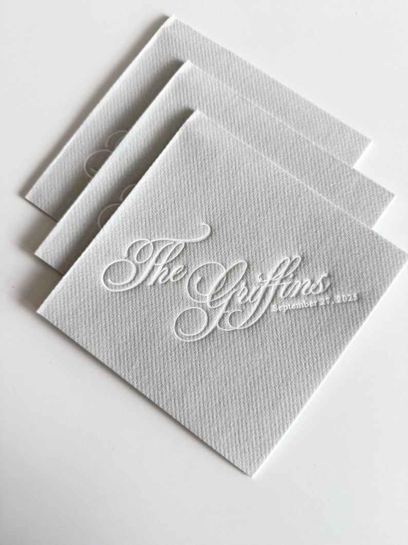 Peut inclure: Une pile de trois serviettes en papier carr&eacute;es gris clair avec une finition textur&eacute;e. La serviette du dessus pr&eacute;sente une calligraphie blanche en relief indiquant The Griffins et la date September 27, 2025, centr&eacute;e sur la surface du papier.