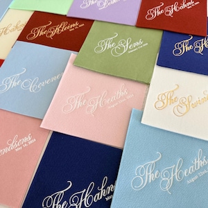 Peut inclure: Une collection de serviettes en papier carr&eacute;es de diff&eacute;rentes couleurs, notamment rouge, bleu, bleu clair, rose, vert et blanc. Chaque serviette pr&eacute;sente un nom et une date personnalis&eacute;s imprim&eacute;s dans une police cursive d&eacute;corative, en blanc ou dor&eacute;.