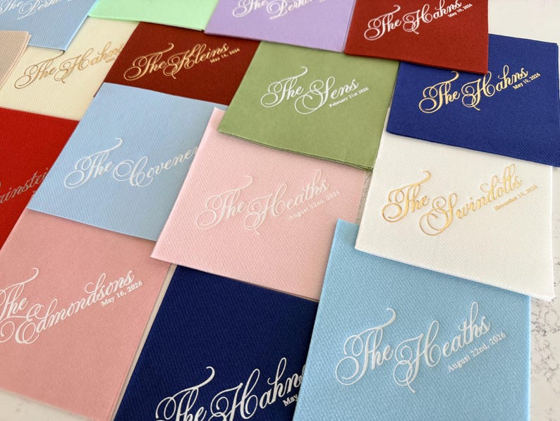 Peut inclure: Une collection de serviettes en papier carr&eacute;es de diff&eacute;rentes couleurs, notamment rouge, bleu, bleu clair, rose, vert et blanc. Chaque serviette pr&eacute;sente un nom et une date personnalis&eacute;s imprim&eacute;s dans une police cursive d&eacute;corative, en blanc ou dor&eacute;.