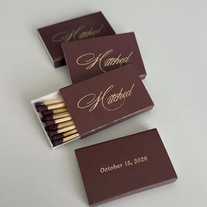 May include: Four rectangular matchboxes in a deep burgundy color. Three boxes feature the word Hitched in gold cursive script. One box displays the text October 15, 2026 in gold print. One box is partially open, revealing wooden matches with dark tips.