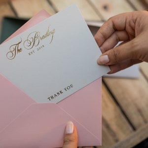 May include: A white card with gold lettering that reads "The Bradley EST 2019" and "THANK YOU" is being held in a pink envelope.