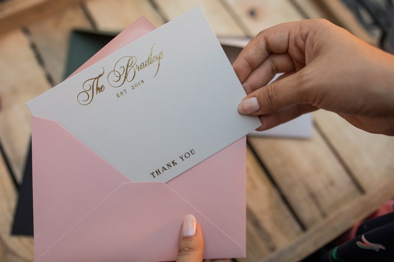 May include: A white card with gold lettering that reads "The Bradley EST 2019" and "THANK YOU" is being held in a pink envelope.