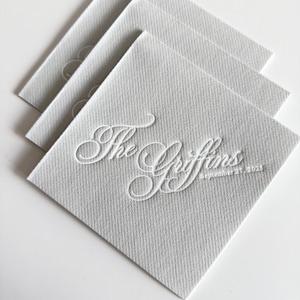 Peut inclure: Une pile de trois serviettes en papier carr&eacute;es gris clair avec une finition textur&eacute;e. La serviette du dessus pr&eacute;sente une calligraphie blanche en relief indiquant The Griffins et la date September 27, 2025, centr&eacute;e sur la surface du papier.