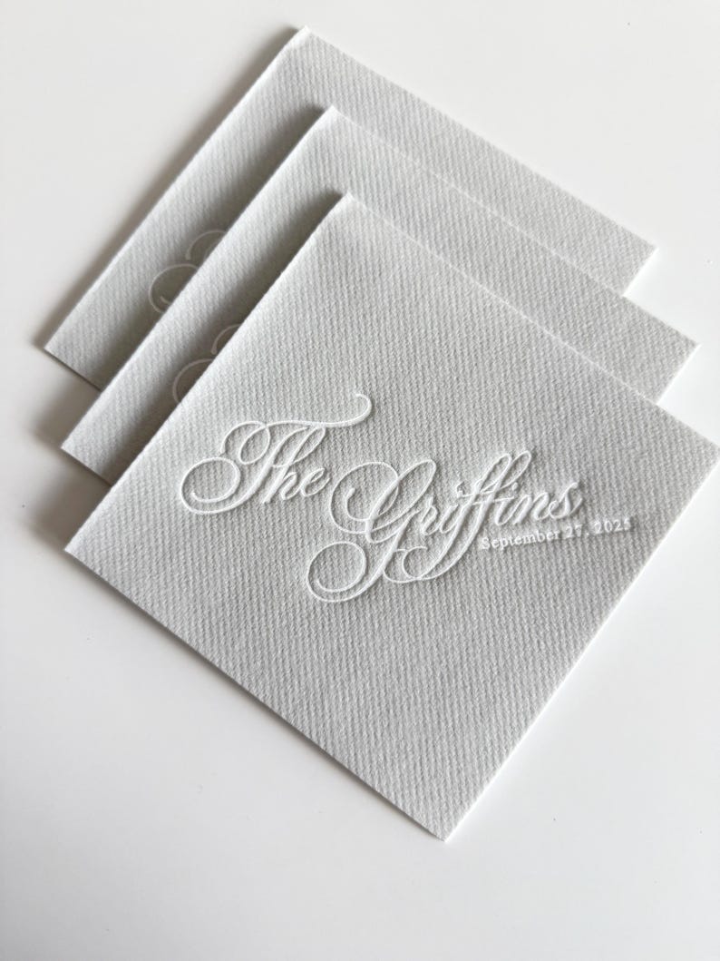 Peut inclure: Une pile de trois serviettes en papier carr&eacute;es gris clair avec une finition textur&eacute;e. La serviette du dessus pr&eacute;sente une calligraphie blanche en relief indiquant The Griffins et la date September 27, 2025, centr&eacute;e sur la surface du papier.