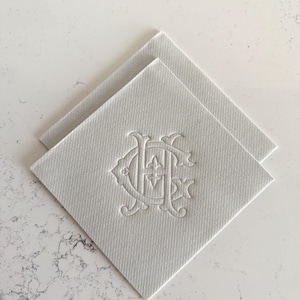 May include: Two white square napkins with an embossed monogram design. The napkins are made of a textured material and are arranged diagonally, showcasing the detailed, elegant pattern. The background is a light, marbled surface.