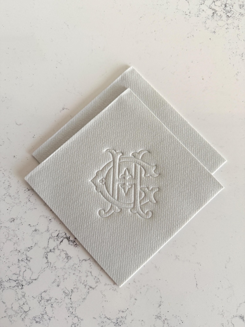 May include: Two white square napkins with an embossed monogram design. The napkins are made of a textured material and are arranged diagonally, showcasing the detailed, elegant pattern. The background is a light, marbled surface.
