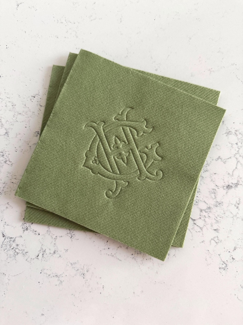 May include: Three olive green square cloth napkins stacked on a white surface. Each napkin features an embossed monogram design in a matching olive green color. The monogram appears to be the letters "A" and "C" intertwined.