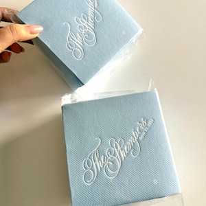 Peut inclure: Deux serviettes carr&eacute;es bleu clair avec une finition textur&eacute;e, chacune emball&eacute;e individuellement dans du plastique transparent. Les serviettes pr&eacute;sentent un texte blanc en relief indiquant The Shempers et la date du 25 avril 2024.