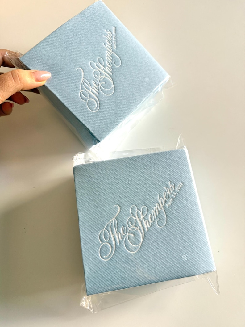 Peut inclure: Deux serviettes carr&eacute;es bleu clair avec une finition textur&eacute;e, chacune emball&eacute;e individuellement dans du plastique transparent. Les serviettes pr&eacute;sentent un texte blanc en relief indiquant The Shempers et la date du 25 avril 2024.