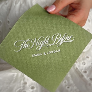 Op de afbeelding: Een vierkant, saliegroen papieren servet met een getextureerde afwerking. De tekst "The Night Before" is gedrukt in wit cursief schrift, met de namen "EMMA & JORDAN" in witte hoofdletters onder het schrift op het oppervlak van het servet.
