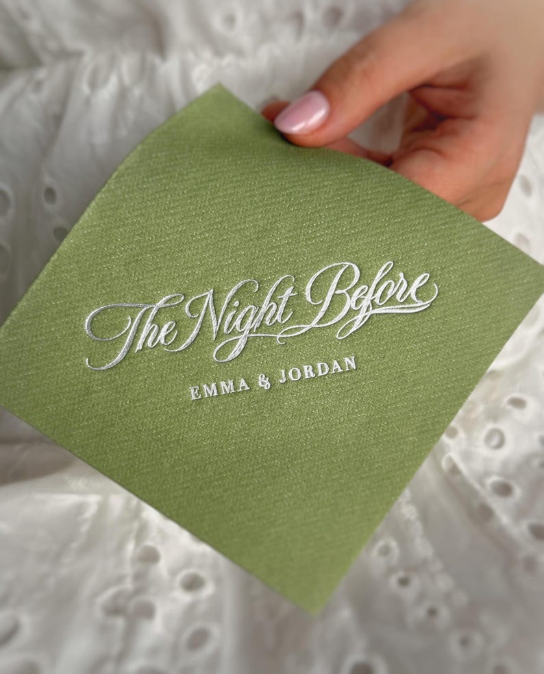 Op de afbeelding: Een vierkant, saliegroen papieren servet met een getextureerde afwerking. De tekst "The Night Before" is gedrukt in wit cursief schrift, met de namen "EMMA & JORDAN" in witte hoofdletters onder het schrift op het oppervlak van het servet.