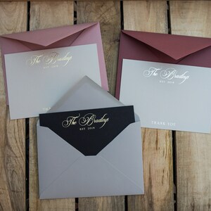 May include: Three envelopes with different colored paper and a black card with gold lettering that reads "The Bradleys Est. 2018". The envelopes are pink, burgundy, and gray. The card is inside the gray envelope.