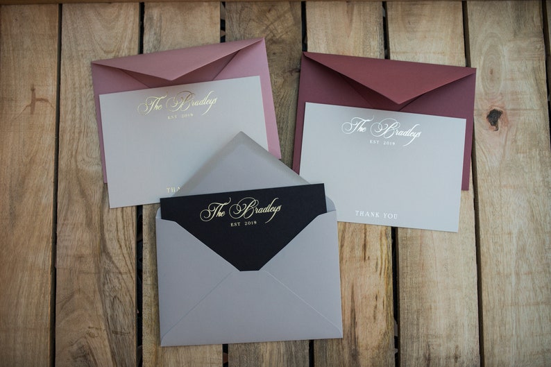 May include: Three envelopes with different colored paper and a black card with gold lettering that reads "The Bradleys Est. 2018". The envelopes are pink, burgundy, and gray. The card is inside the gray envelope.
