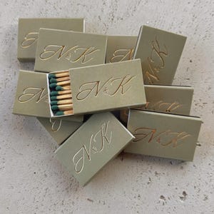 Könnte beinhalten: Eine Ansammlung olivgrüner Streichholzschachteln, jede mit den Initialen "NK" in goldener Schrift geprägt. Eine Schachtel ist geöffnet und zeigt Streichhölzer mit grünen Köpfen. Die Schachteln liegen auf einer strukturierten, hellgrauen Oberfläche.