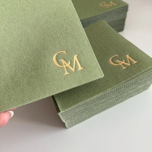 以下が含まれることがあります： GMの文字がゴールドの箔押しで刻印された、正方形のセージグリーンの紙ナプキンの束。質感のある紙はマットな仕上がりで、メタリックゴールドのロゴが各ナプキンの前面中央に配置されています。