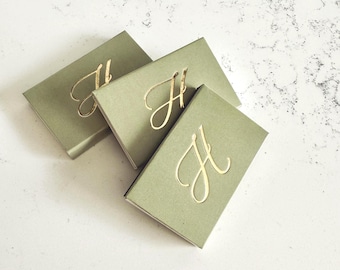 May include: Three rectangular sage green boxes, each featuring a gold foil embossed letter H in a cursive script font centered on the lid. The boxes are arranged in a slightly overlapping cluster, showcasing the matte finish and metallic lettering.