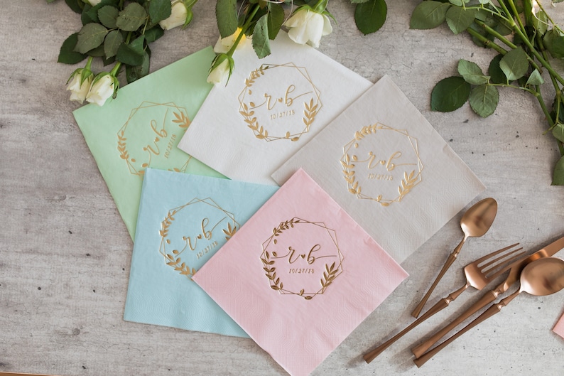 Pu&ograve; includere: Cinque tovaglioli di diversi colori con lettere dorate. I tovaglioli sono piegati a met&agrave; e hanno una cornice geometrica dorata con un motivo floreale e le iniziali "r & b" e la data "10.21.2023".