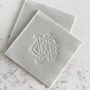 May include: Two white square coasters with a textured surface. One coaster is on top of the other. Each coaster has an embossed monogram design in the lower right corner. The coasters are on a white and gray marble surface.