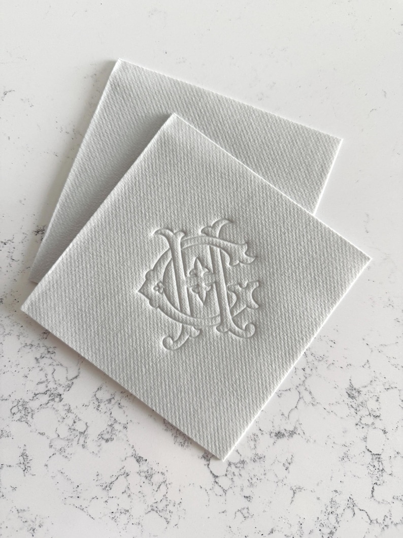 May include: Two white square coasters with a textured surface. One coaster is on top of the other. Each coaster has an embossed monogram design in the lower right corner. The coasters are on a white and gray marble surface.