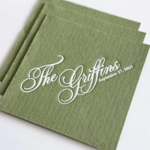 Peut inclure: Une pile de serviettes en papier carr&eacute;es vert sauge avec un texte blanc en relief indiquant The Griffins et la date du 27 septembre 2025. Les serviettes pr&eacute;sentent une finition textur&eacute;e semblable &agrave; du lin et sont dispos&eacute;es en quinconce.