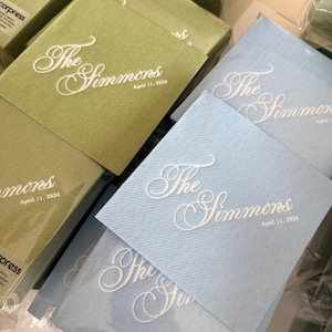 Peut inclure: Piles de serviettes en papier carr&eacute;es vert sauge et bleu clair. Chaque serviette pr&eacute;sente un texte blanc en relief indiquant The Simmons et la date du 11 avril 2026. Les serviettes sont emball&eacute;es dans du plastique transparent avec une &eacute;tiquette decorpress.