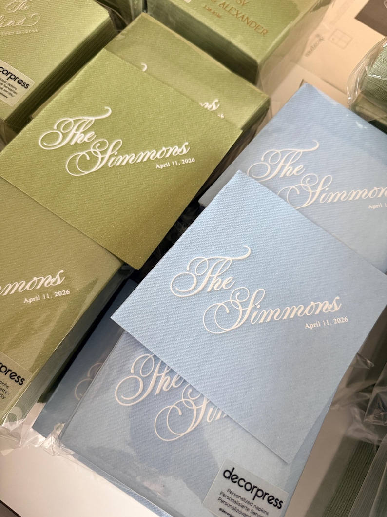 Peut inclure: Piles de serviettes en papier carr&eacute;es vert sauge et bleu clair. Chaque serviette pr&eacute;sente un texte blanc en relief indiquant The Simmons et la date du 11 avril 2026. Les serviettes sont emball&eacute;es dans du plastique transparent avec une &eacute;tiquette decorpress.