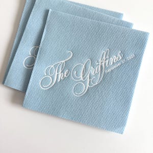 Peut inclure: Une pile de serviettes en papier carr&eacute;es bleu clair avec un texte blanc en relief indiquant The Griffins et la date du 27 septembre 2025 dans une police cursive. Les serviettes pr&eacute;sentent une surface textur&eacute;e et sont l&eacute;g&egrave;rement superpos&eacute;es.