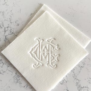 May include: Two off-white square napkins with a textured surface. Each napkin features an embossed monogram design in the center. The monogram appears to be two intertwined letters, possibly initials, with decorative flourishes.