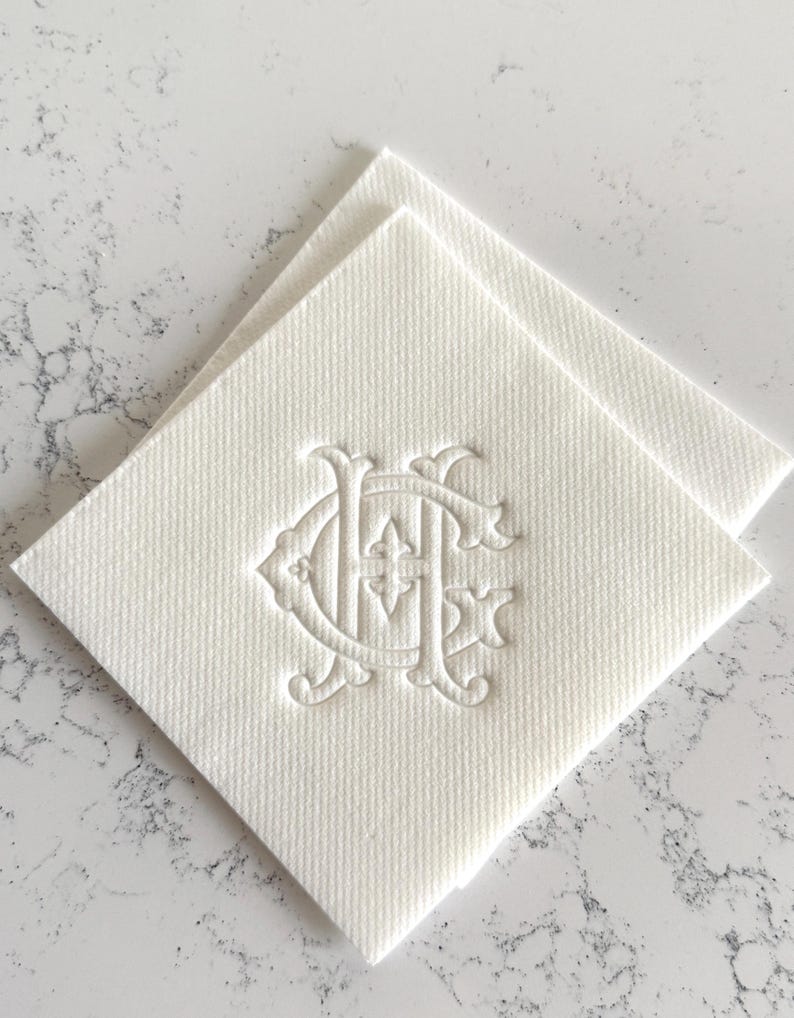 May include: Two off-white square napkins with a textured surface. Each napkin features an embossed monogram design in the center. The monogram appears to be two intertwined letters, possibly initials, with decorative flourishes.