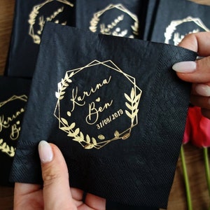 Pu&ograve; includere: Tovaglioli neri con lettering dorato. I tovaglioli presentano una cornice geometrica con una ghirlanda floreale e il testo "Karina & Ben 31/08/2019".