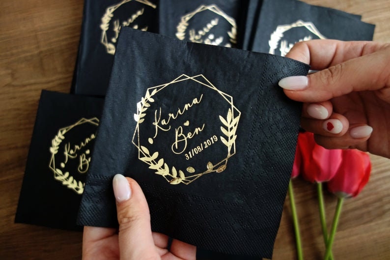 Pu&ograve; includere: Tovaglioli neri con lettering dorato. I tovaglioli presentano una cornice geometrica con una ghirlanda floreale e il testo "Karina & Ben 31/08/2019".