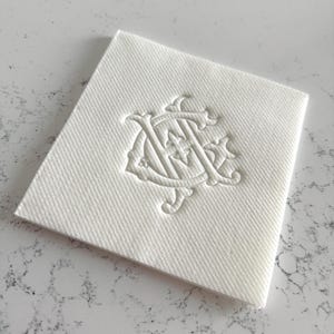May include: A square, white linen napkin with a textured surface. The napkin features an embossed monogram design in the center. The monogram is an ornate, intertwined design, likely for formal events or personalized use.