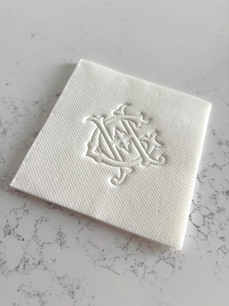 May include: A square, white linen napkin with a textured surface. The napkin features an embossed monogram design in the center. The monogram is an ornate, intertwined design, likely for formal events or personalized use.